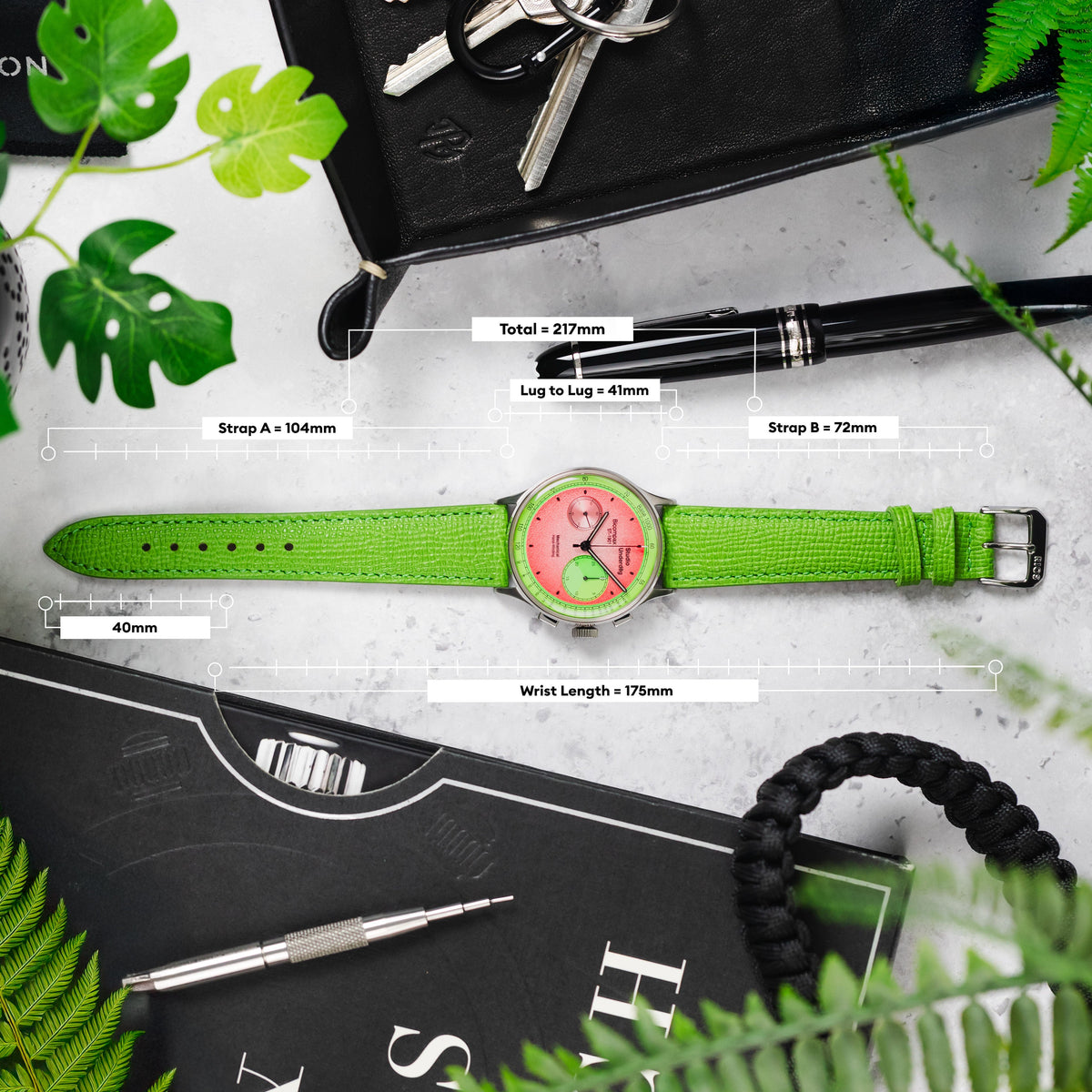 Image showing the measurements of a green watch strap and pink/green watch, with labels for total length, lug width, strap lengths, and wrist length. The watch is arranged with other items like keys, a pen, and a book.