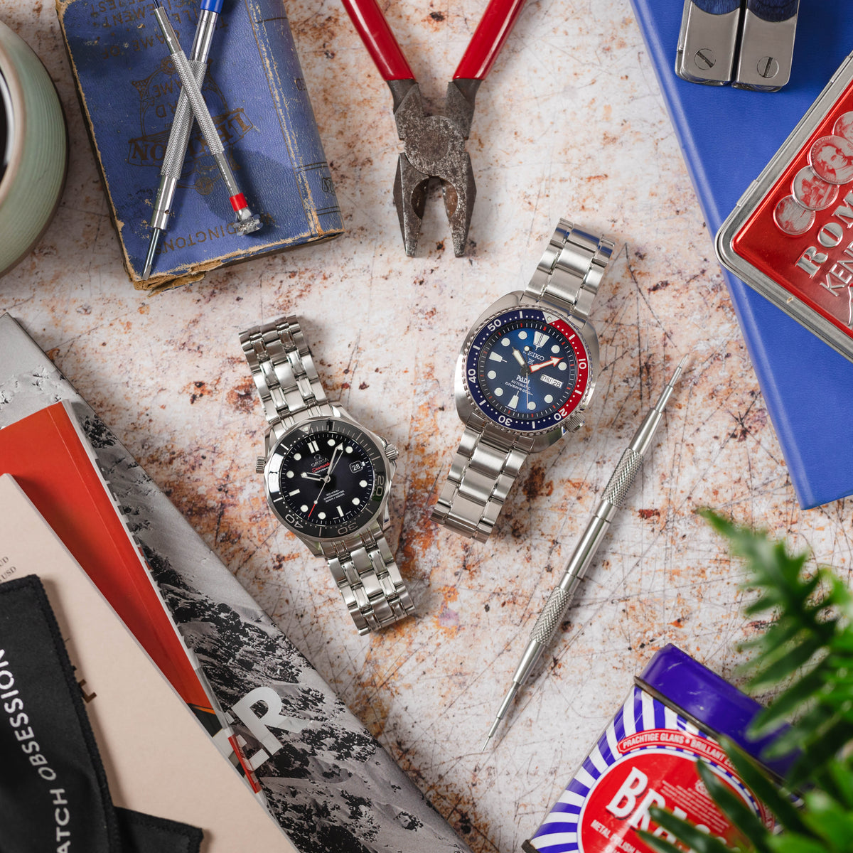 A top-down shot of two watches with metal bracelets placed on a rustic, worn metal surface. The watch on the left has a black dial, and the one on the right has a blue and red bezel with a blue dial. Surrounding the watches are various objects, including