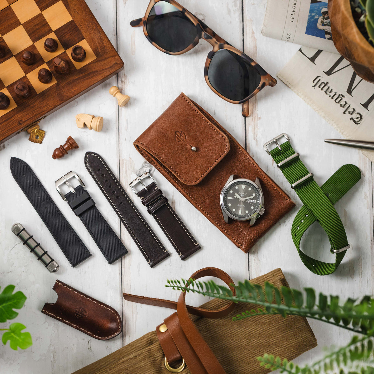 A flat-lay shot of a variety of watch accessories and straps on a white wooden surface. The image includes a Seiko 5 Sports watch in a leather watch pouch, a selection of different watch straps, a chessboard, and other items such as sunglasses, a newspap
