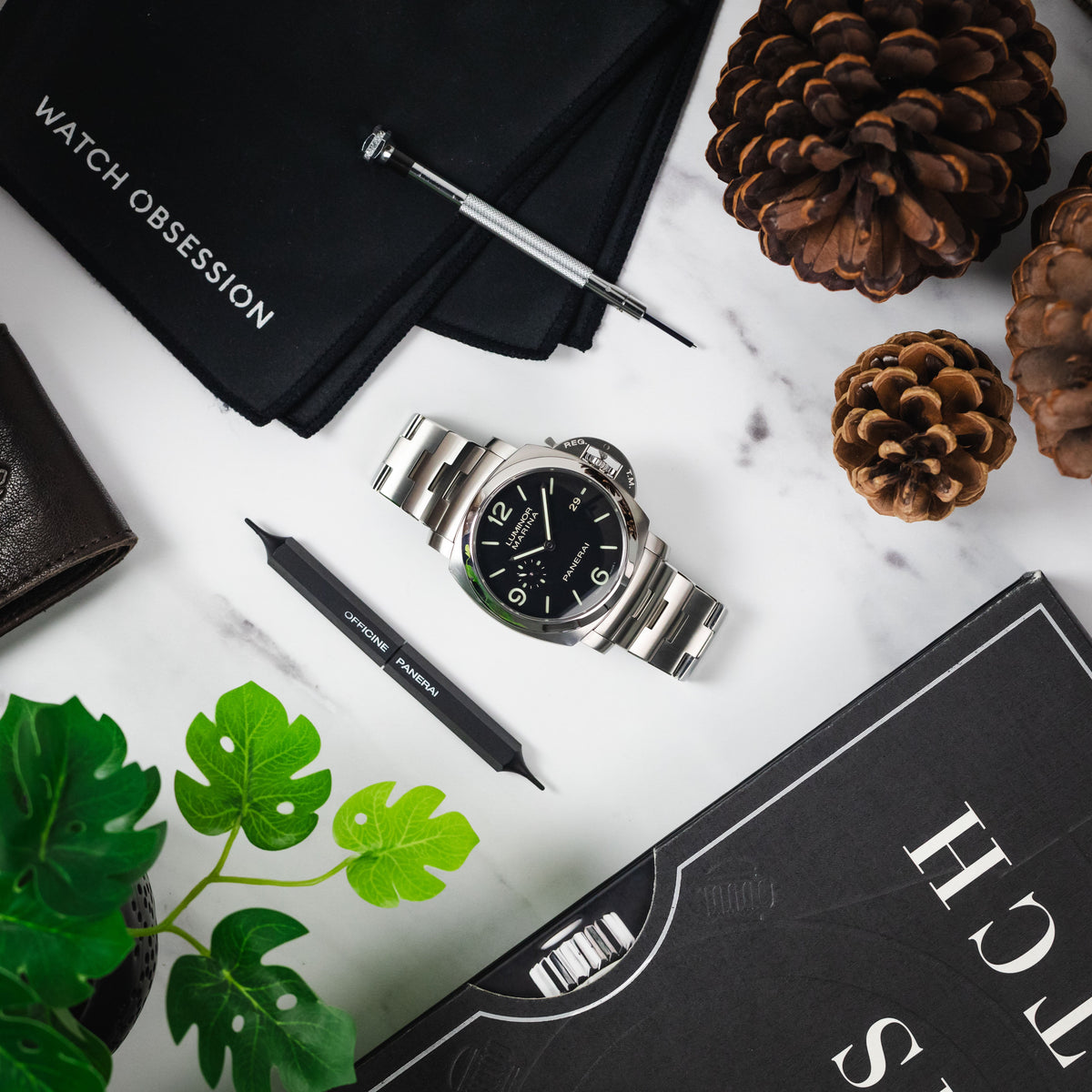 Overhead view of a Panerai Luminor Marina on a steel bracelet, with a watch cloth, tool, pen, book, plant, & pine cones, arranged in a still life.
