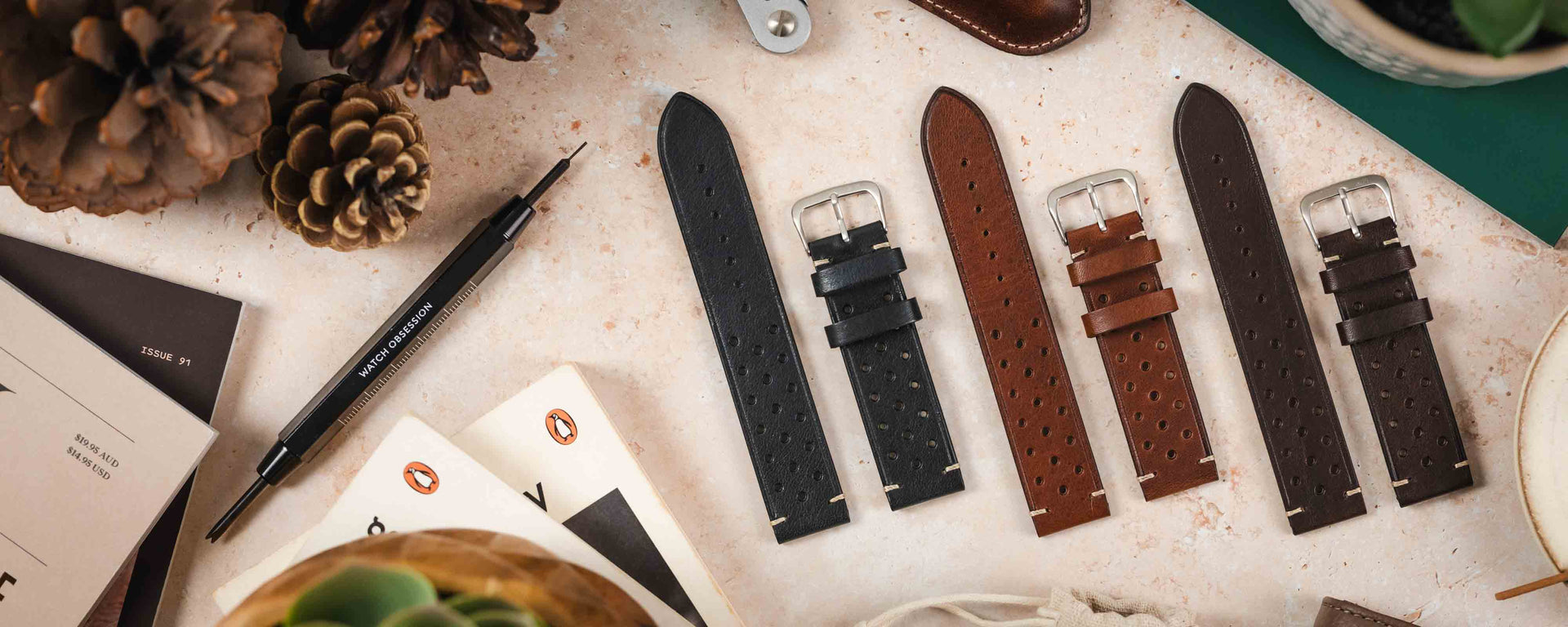 A flat-lay photograph showing three Brahms watch straps—black, brown, and dark brown—arranged on a light-colored surface with pine cones, a spring bar tool, and paperback books.