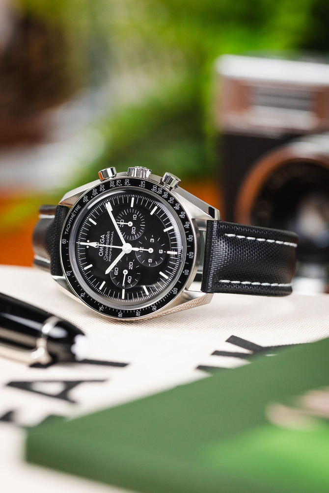 Curved black Artem sailcloth watch strap with wite stitching fitted to black Omega Speedmaster Moonwatch on a table. 