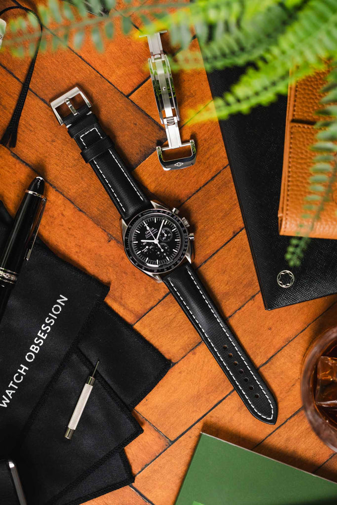 High angle shot of Black Omega Speedmaster Moonwatch fitted with Artem Straps  black sailcloth classic watch strap with white stitching and stanless steel artem logo embossed tangle buckle on wooden table featuring Artem Straps Loop-less buckle. 