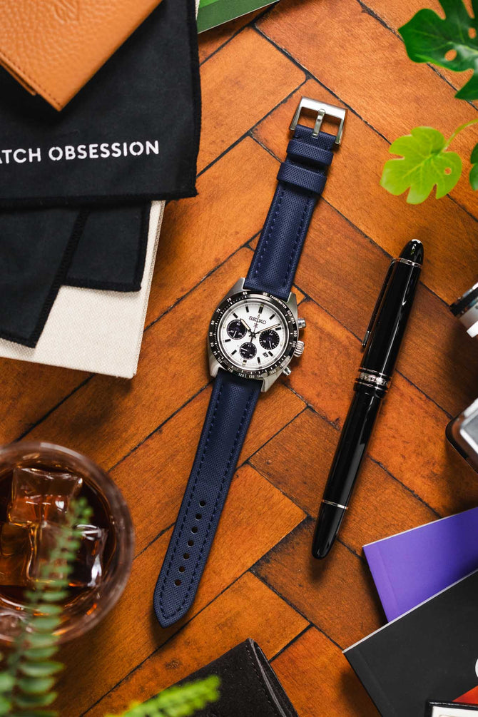 Seiko Speedtimer timepiece with Panda Chronograph with a Navy Blue Artem Straps Classic Sailcloth watch strap with Silver pin buckle next to a black pen on top of wooden table. 