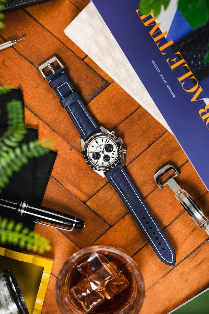 Seiko Speedtimer timepiece with Panda Chronograph with a Navy Blue Artem Straps Classic Sailcloth with white stitching watch strap and Silver pin buckle next to a black pen and Artem Loop-less clasp on top of wooden table.