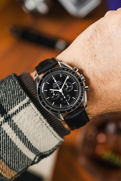 Black Sailcloth watch strap with black stitching fitted to a Omega Speedmaster Time watch with a black dial  and silver  pusher and crown, on a wrist with check shirt with black and cream.