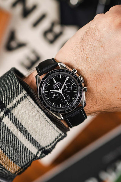 Black Sailcloth watch strap with grey stitching fitted to a Omega Speedmaster Moonwatch Time Piece with a black dial and silver pusher and crown, on a wrist with check shirt with black and cream.