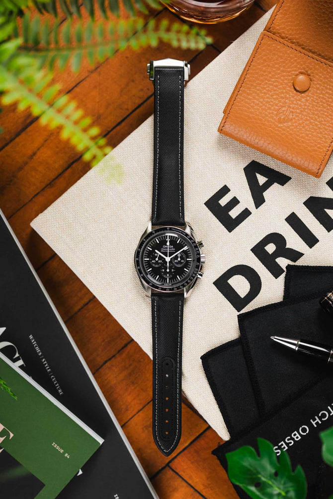 Artem Straps Loop-Less Black Sailcloth with grey stitching watch strap fitted to a black Omega Speedmaster Moonwatch timepiece on a wooden table. 