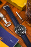 Navy Blue Artem Loop-Less sailcloth watch strap fitted to Black Omega Speedmaster Moonwatch face up on woooden desk next to open Artem Loop-Less deployment clasp