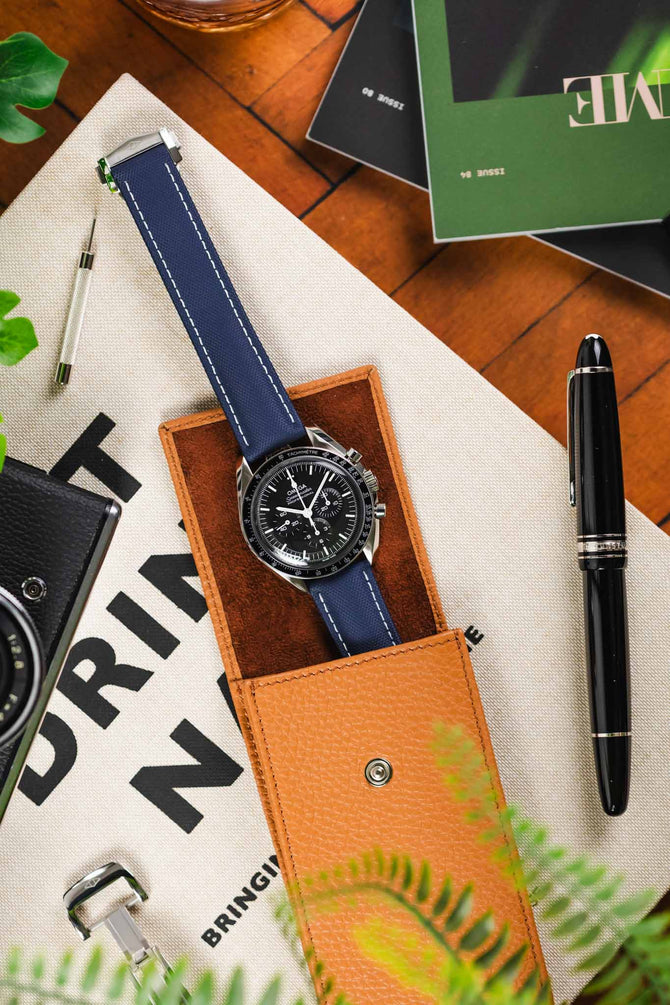 Black Omega Speedmaster moonwatch fitted to Navy Blue with white stitching Artem Straps Loop-less sailcloth watch strap in beige leather watch case next to black pen on wooden table
