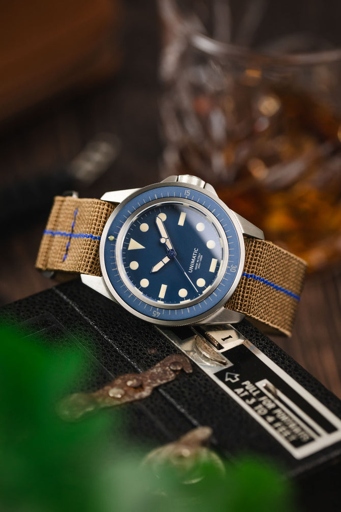 Blue dial watch leant on a vintage camera fitted to a brown parachute webbing watch strap with brushed stainless steel hardware