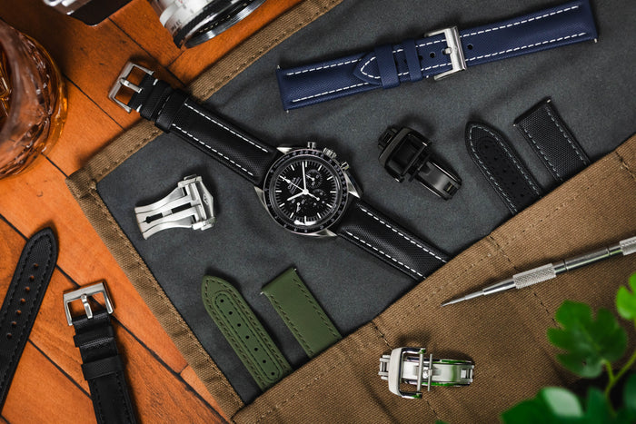 An image of an Omega Speedmaster lay flat on an open watch roll attached to a black Artem sailcloth strap, surrounded by Artem straps and clasps against a wooden background.