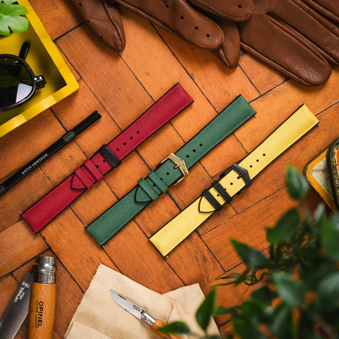 An overhead flat lay featuring three Hirsch Arne sailcloth effect watch straps in red, green, and yellow, each with a contrasting buckle. They are artfully arranged on a warm wooden background alongside brown leather driving gloves, dark sunglasses in a y