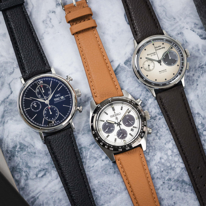 An overhead view of three watches with leather straps on a grey and white marbled surface: an automatic dark blue dial IWC, a white dial Quartz Seiko chronograph, and a cream dial Mechanical Studio Underd0g.