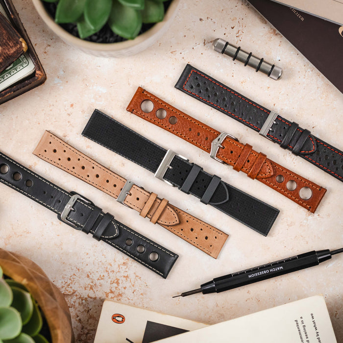 A top-down flat lay features an assortment of watch straps laid out on a light, textured surface. From left to right, there's a black rally strap with white stitching, a tan rally strap, a black rubber rally strap, a brown rally strap, and a black rally s