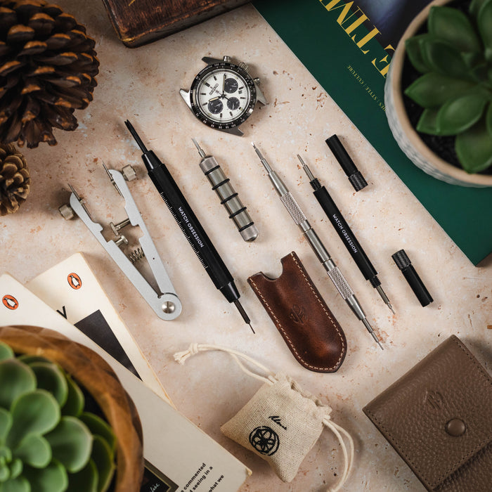  A top-down flat lay displays a collection of watch tools and accessories on a light, textured surface. Included are a spring bar tool, a watch case opener, various screwdrivers, a small leather pouch, and different size spring bars. The arrangement also 