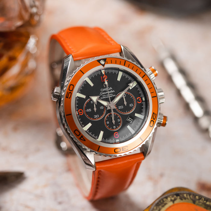 A detailed shot of an Omega Seamaster Planet Ocean Chronograph watch, model 2918.50.83, featuring a black dial, an orange unidirectional bezel, and an orange rubber strap, resting on a textured light-colored surface.