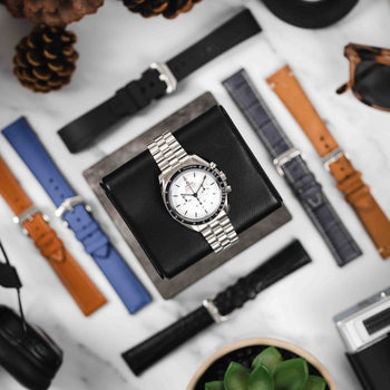 Assorted custom watch straps in black, blue, and tan surrounding an Omega Speedmaster Professional watch with a steel bracelet. Enhance your timepiece.