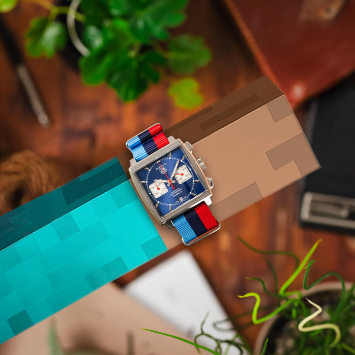 Close-up of a TAG Heuer Monaco watch with a blue dial on a blocky Minecraft arm. Wooden background.