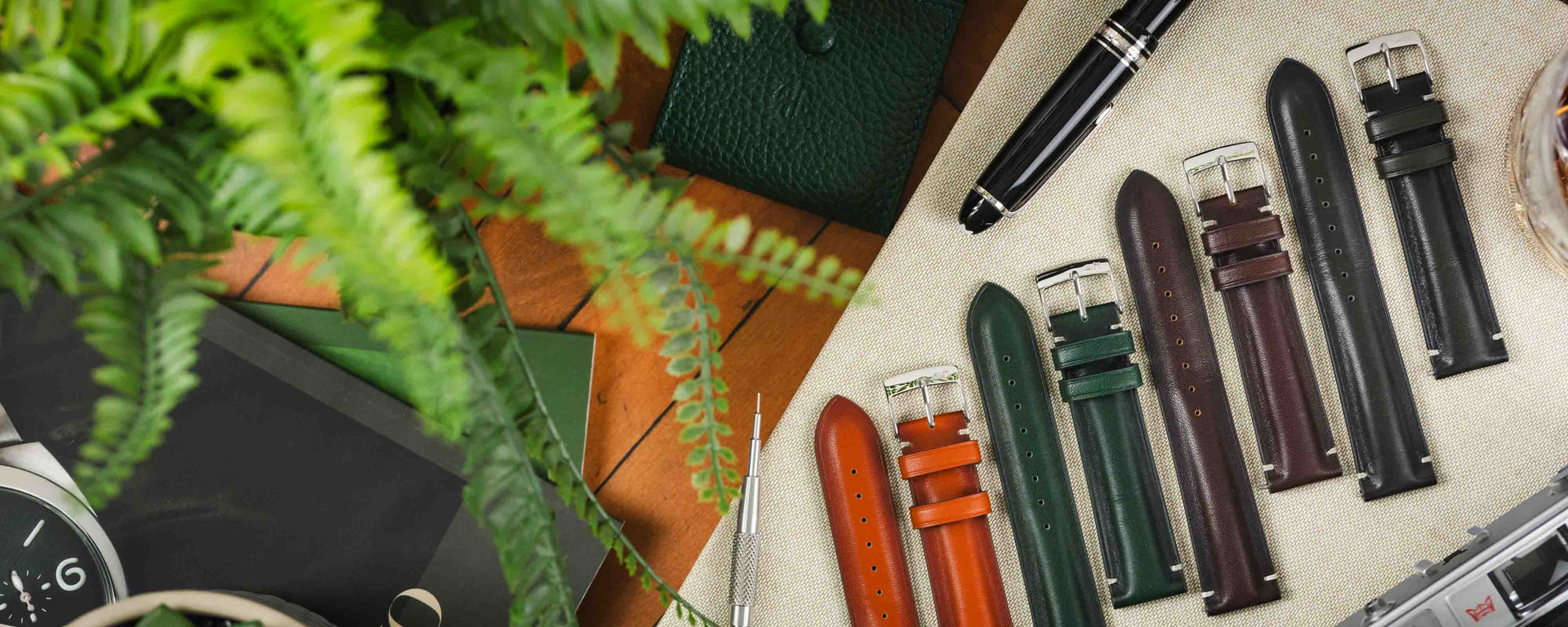 A flat-lay photograph showing three Brahms watch straps—black, brown, and dark brown—arranged on a light-colored surface with pine cones, a spring bar tool, and paperback books.
