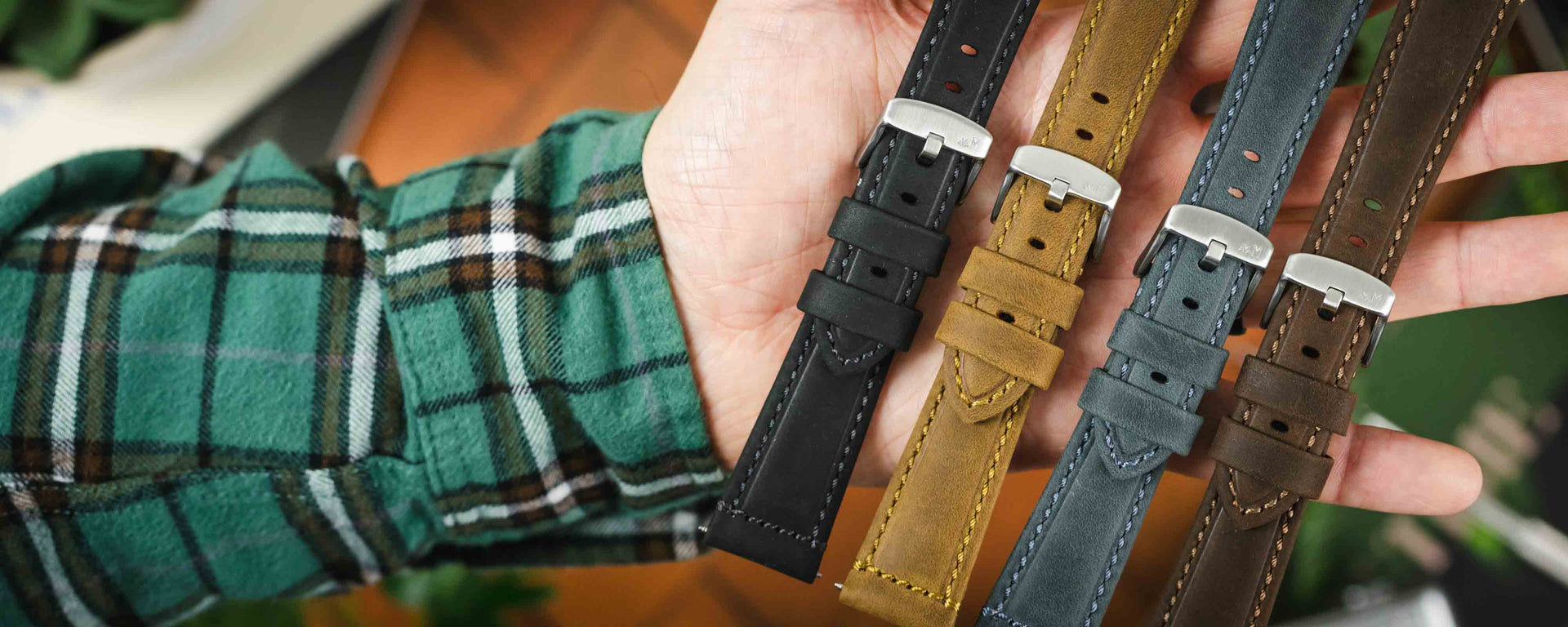  A flat-lay photograph showing three Brahms watch straps—black, brown, and dark brown—arranged on a light-colored surface with pine cones, a spring bar tool, and paperback books.