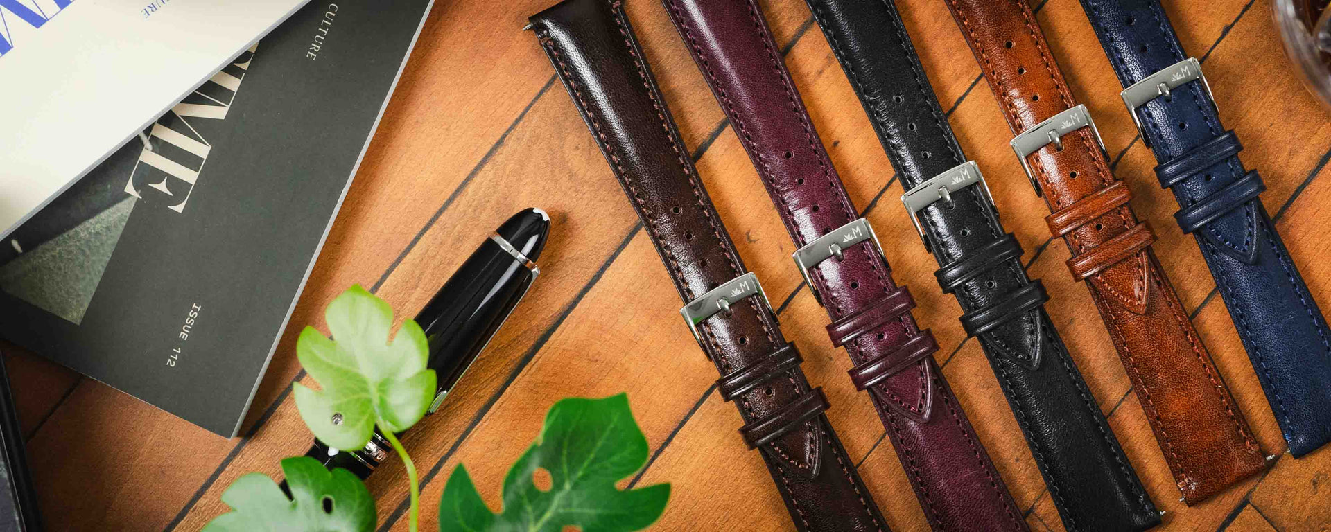 A flat-lay photograph showing three Brahms watch straps—black, brown, and dark brown—arranged on a light-colored surface with pine cones, a spring bar tool, and paperback books.