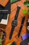 Artem Loop-Less Black Sailcloth Watch Strap  and Artem Straps logo embossed Loop-Less Deployment Clasp fitted to Black Omega Speedmaster faceup on a wooden table next to black pen