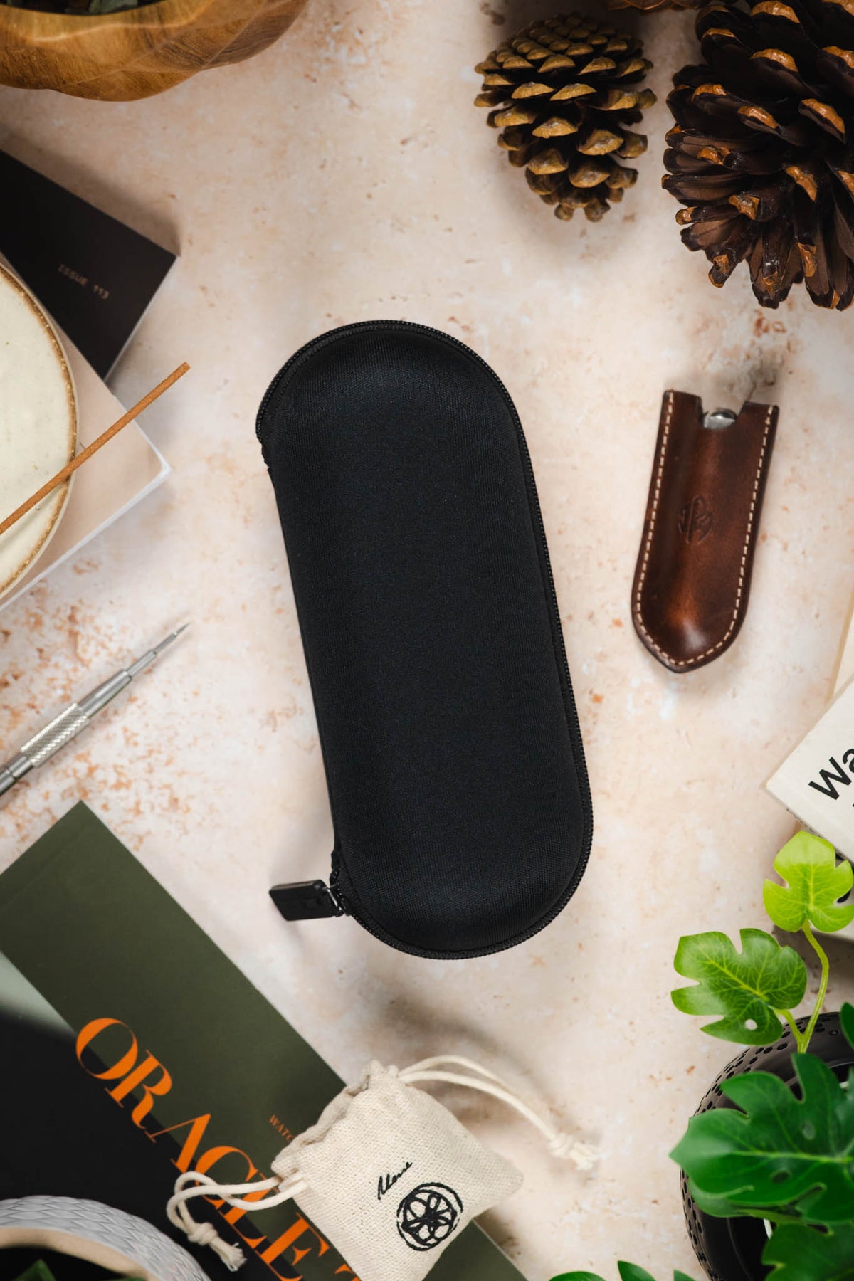 Black hard-shell watch travel case with a textured fabric exterior and zip-around closure, displayed flat on a tabletop surrounded by watch accessories and decorative items.