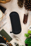 Black hard-shell watch travel case with a textured fabric exterior and zip-around closure, displayed flat on a tabletop surrounded by watch accessories and decorative items.