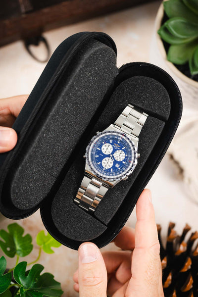 Black zippered watch travel case opened to reveal a luxury stainless steel wristwatch secured in a protective foam-lined interior for safe storage and transport.