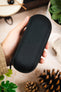 Black hard-shell zippered watch storage case with a textured fabric exterior, held in hand against a decorative tabletop background with plants and pinecones.