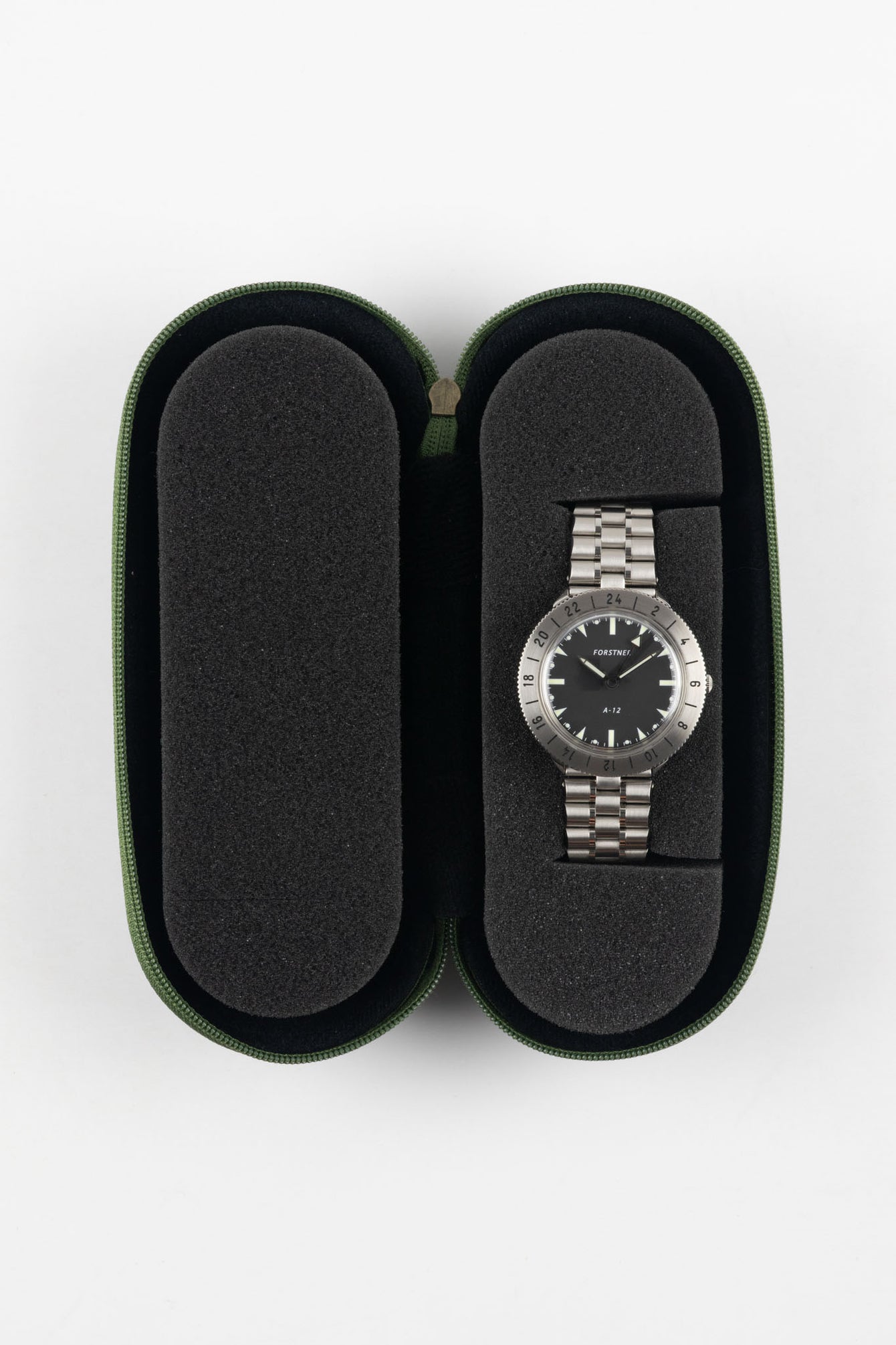 Open green zippered watch case with a black foam interior, featuring two padded compartments; a stainless steel bracelet watch with a black dial is secured in the right slot, while the left slot is empty, set against a plain light background.