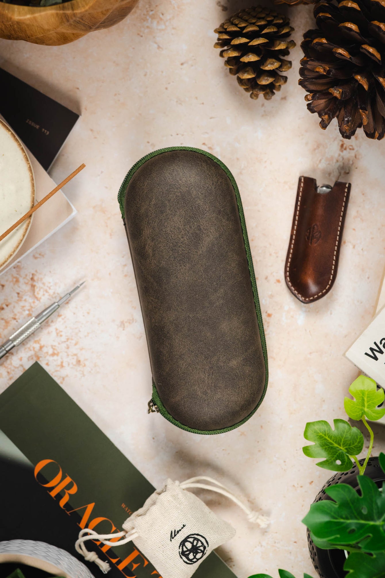 Green hard-shell watch travel case with a distressed vintage-look exterior and matching green zip-around closure, displayed on a tabletop alongside watchmaking tools and accessories.