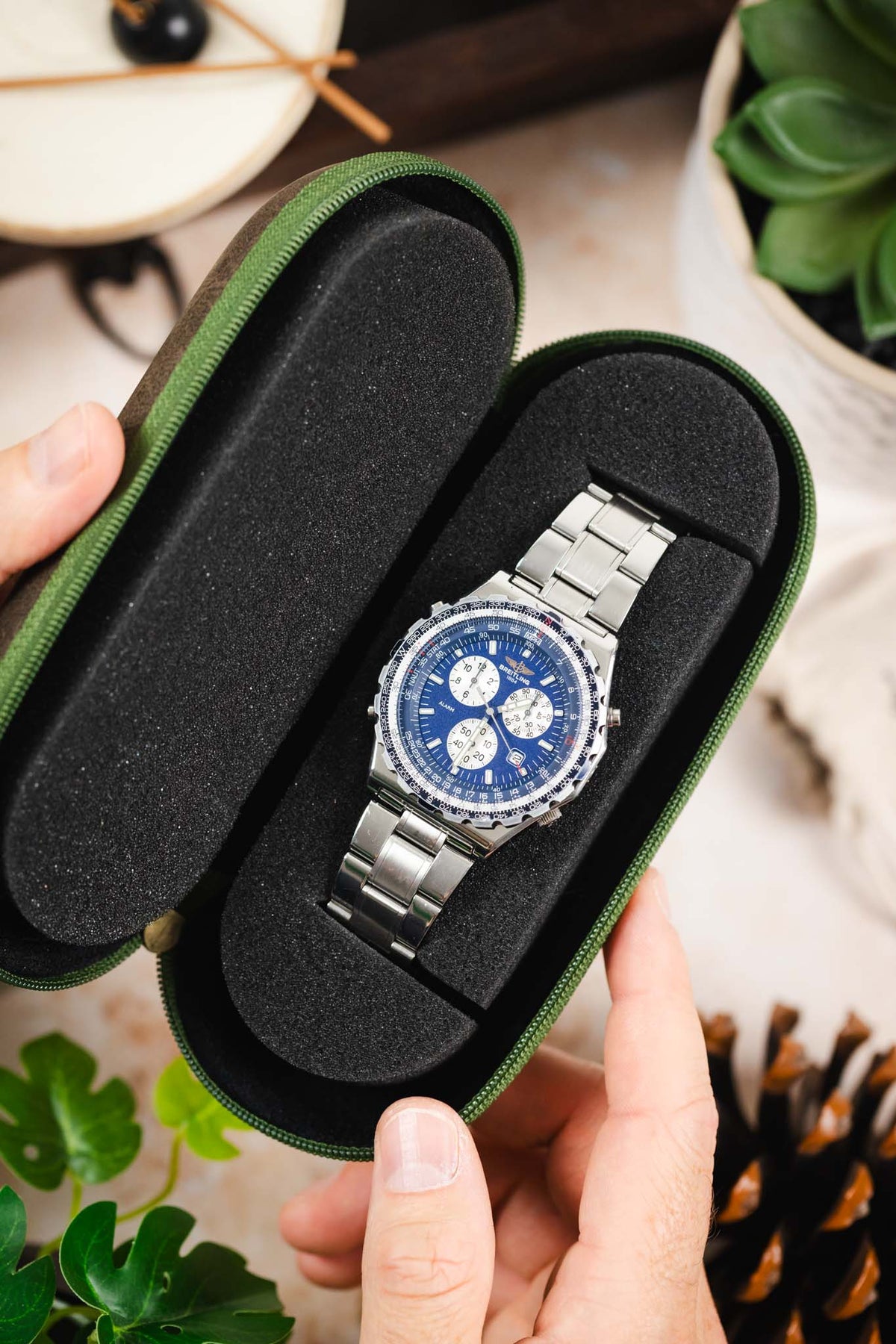 Open green hard-shell watch travel case with a foam-lined interior, displaying a stainless steel wristwatch securely positioned on a padded insert for protection during storage and travel.