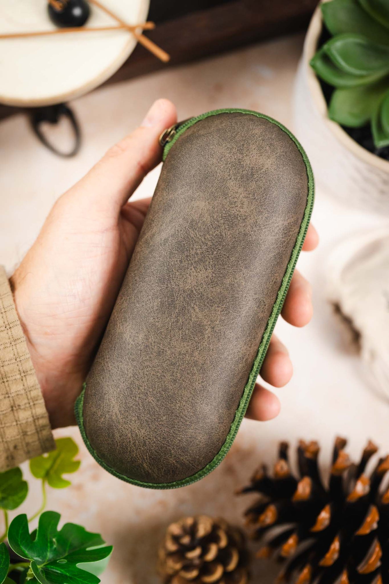 Green hard-shell watch travel case with a distressed fabric exterior and matching green zip-around closure, held in hand against a decorative tabletop background.