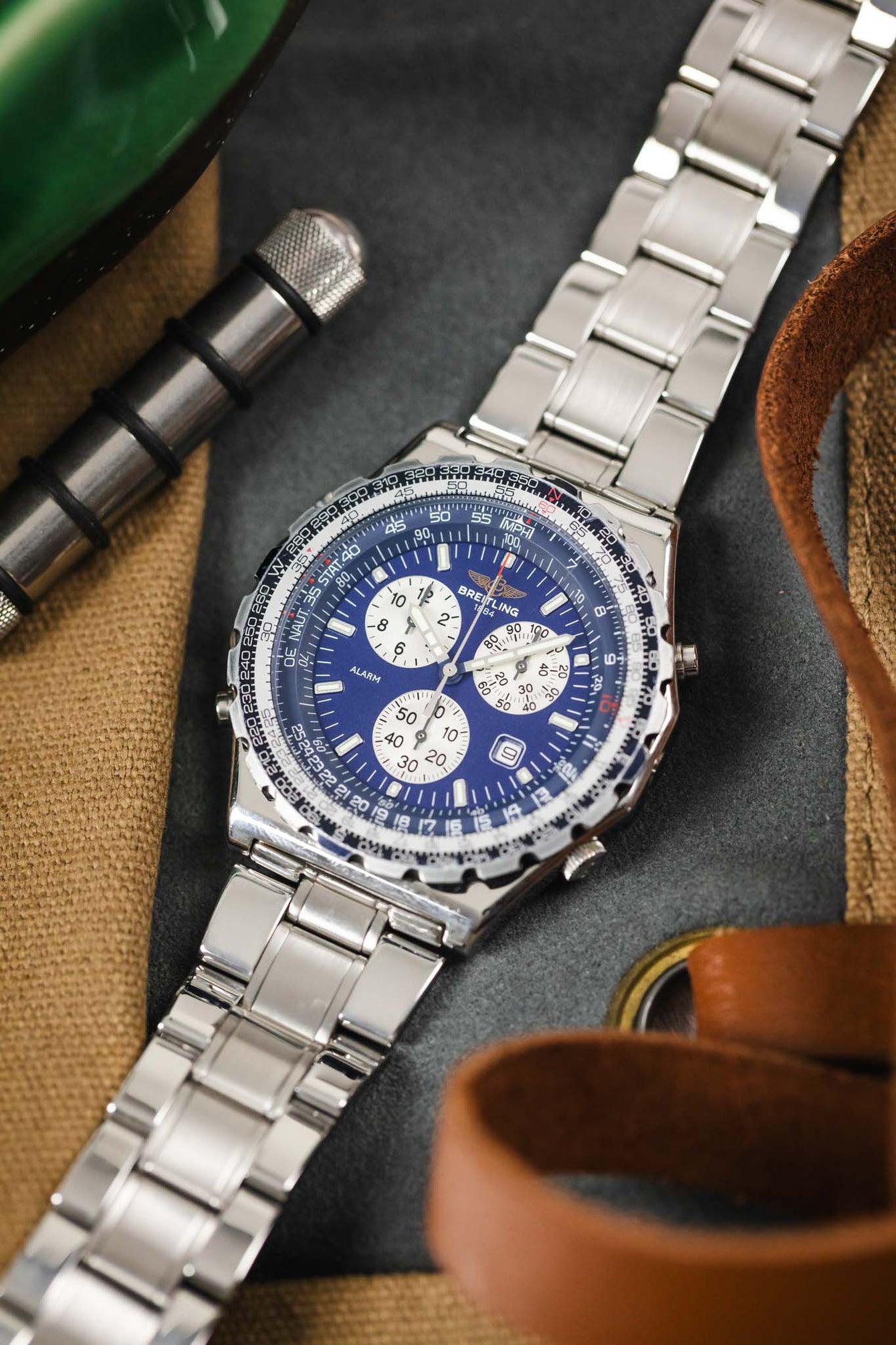 A top-down lifestyle shot of a Breitling Jupiter Pilot watch laying on a grey and tan surface next to a metal spring bar tool and leather straps.