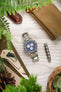 An overhead lifestyle photograph of a Breitling Jupiter Pilot watch centered on a light wood grain surface, framed by green fern leaves and travel accessories.