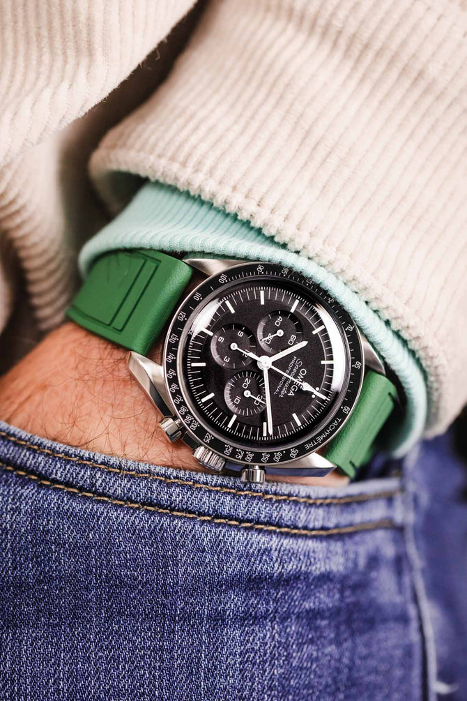 Pocket shot of a Speedmaster in jeans pocket with turquoise hoodie and cream cord shirt, the watch is fitted to a green rubber strap