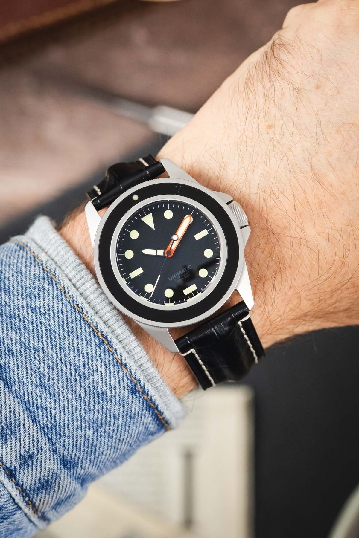 Unimatic dive watch on a black alligator-grain leather strap with cream contrast stitching, shown on wrist with a black dial and bezel.