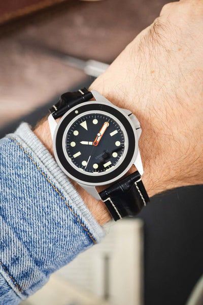 Unimatic dive watch on a black alligator-grain leather strap with cream contrast stitching, shown on wrist with a black dial and bezel.