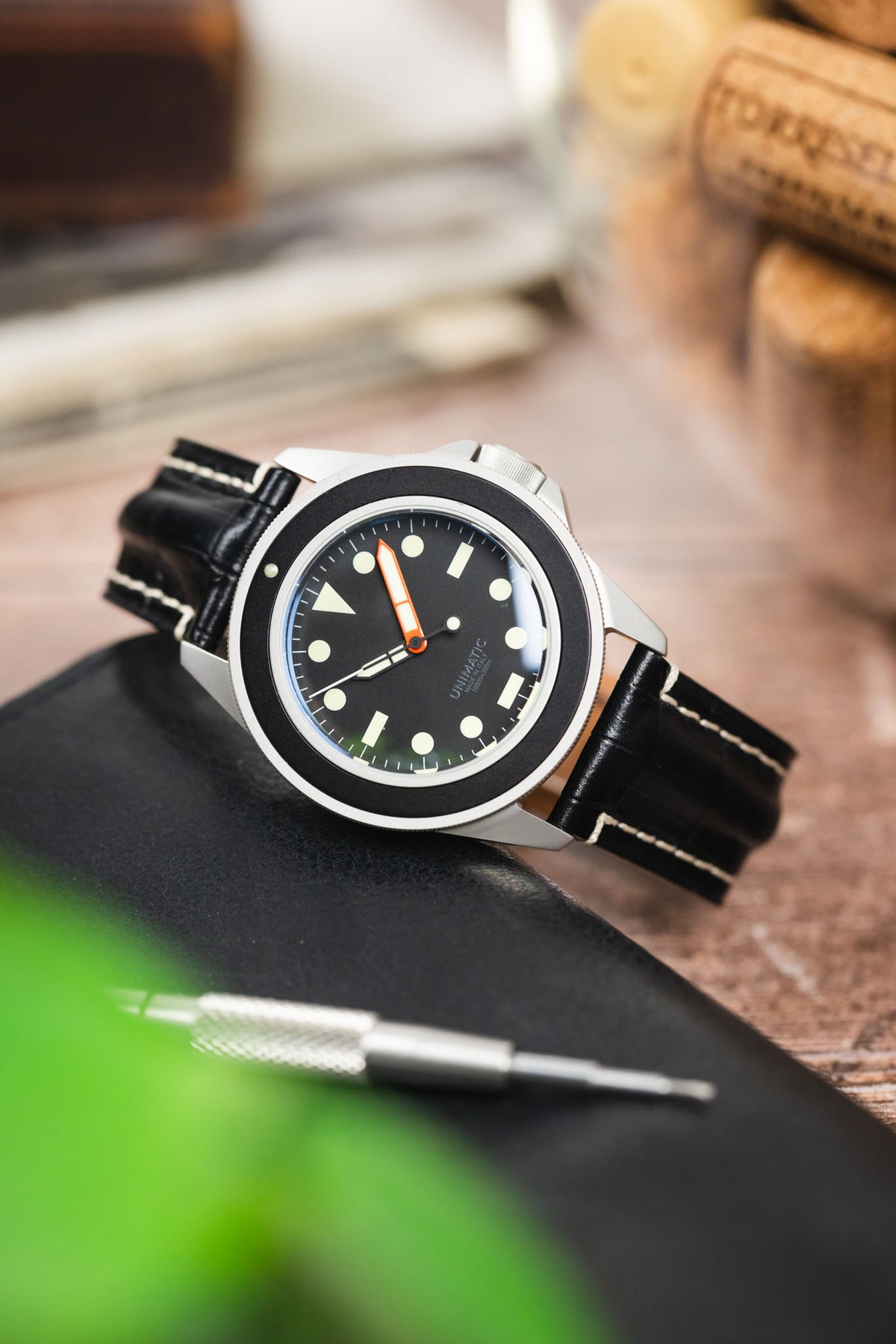 Unimatic dive watch on a black alligator-grain leather strap with cream contrast stitching, displayed on a leather notebook with watchmaking tools and vintage accessories.