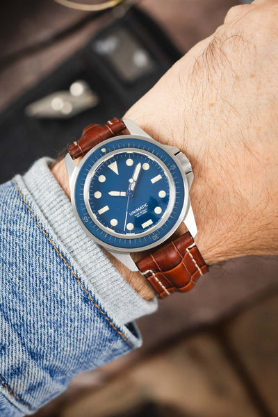 Unimatic dive watch on a gold brown alligator-grain leather strap with cream contrast stitching, shown on wrist with a blue dial and bezel.