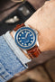 Unimatic dive watch on a gold brown alligator-grain leather strap with cream contrast stitching, shown on wrist with a blue dial and bezel.