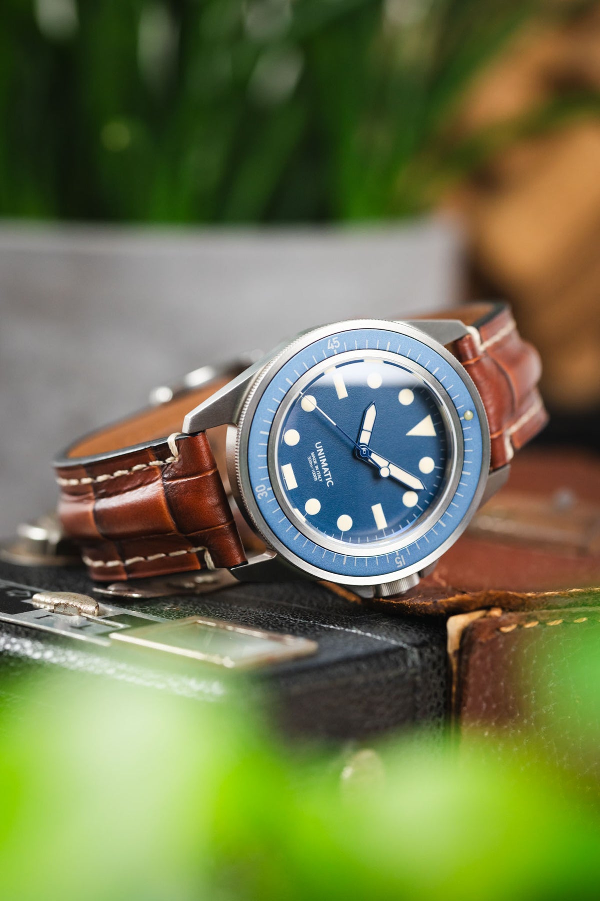 Unimatic dive watch on a gold brown alligator-grain leather strap with cream contrast stitching, displayed on vintage camera equipment with a blue dial and bezel.