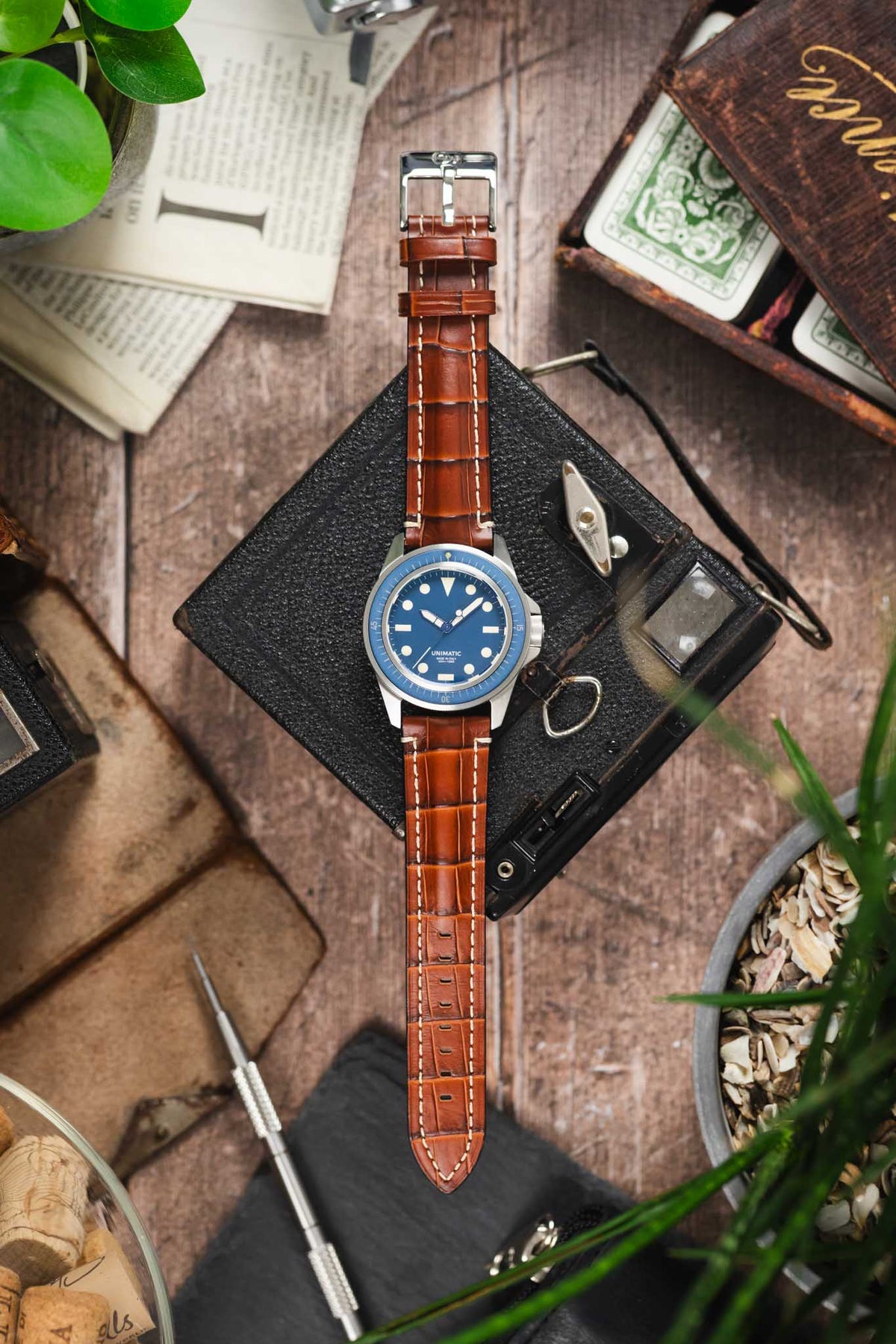 Unimatic dive watch on a gold brown alligator-grain leather strap with cream contrast stitching, displayed with vintage accessories on a rustic wooden surface.