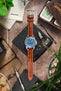 Unimatic dive watch on a gold brown alligator-grain leather strap with cream contrast stitching, displayed with vintage accessories on a rustic wooden surface.