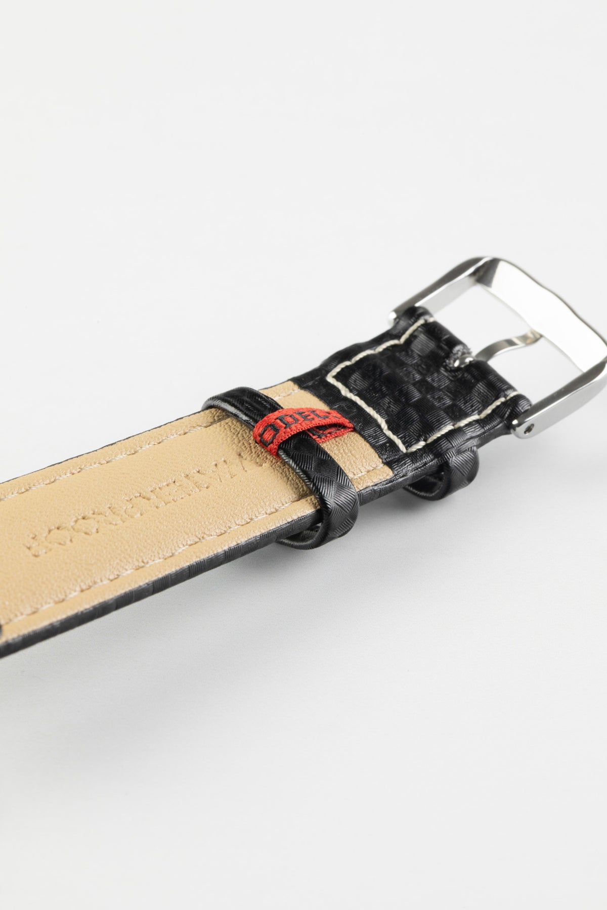 Black carbon-fibre-pattern leather watch strap with a beige leather lining, red branded tab, and polished stainless steel buckle, shown in close-up on a white background.