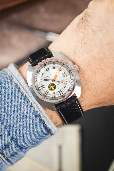 Doxa SUB 300 dive watch on a black carbon-fibre-pattern leather strap with white contrast stitching, shown on wrist with a white dial and orange minute hand.