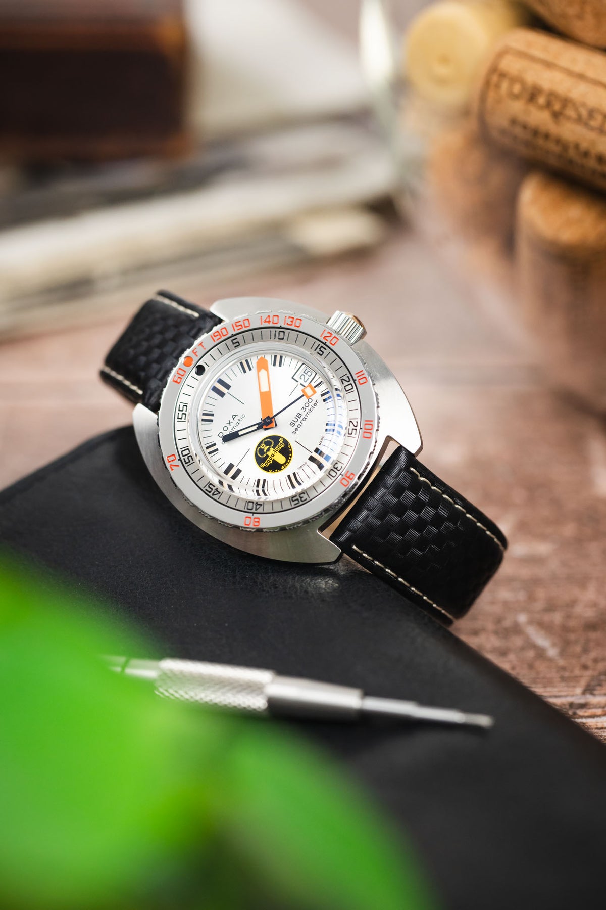 Doxa SUB 300T dive watch on a black carbon-fibre-pattern leather strap with white contrast stitching, displayed on a leather notebook with watchmaking tools and vintage accessories.