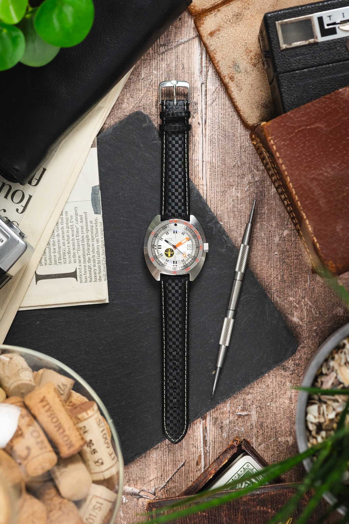 Doxa dive watch on a black carbon-fibre-pattern leather strap with white contrast stitching, displayed with vintage accessories on a rustic wooden surface.